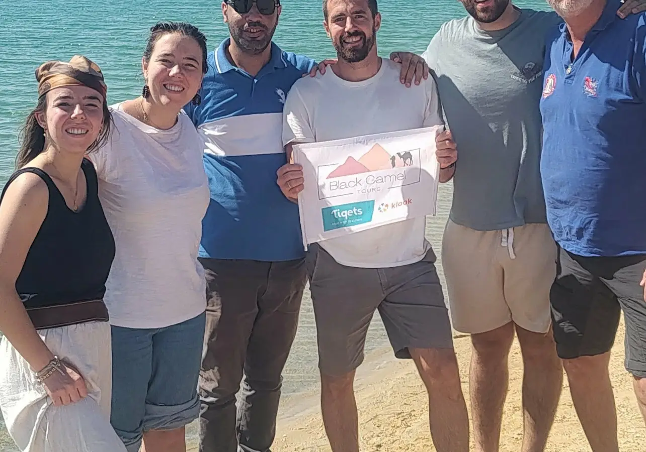 Six people stand together on a sandy beach by a lake, with two holding a sign that reads "Black Camp Tigets."