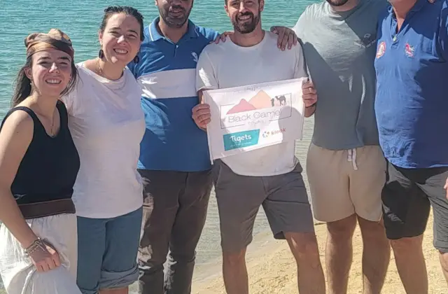 Six people stand together on a sandy beach by a lake, with two holding a sign that reads "Black Camp Tigets."