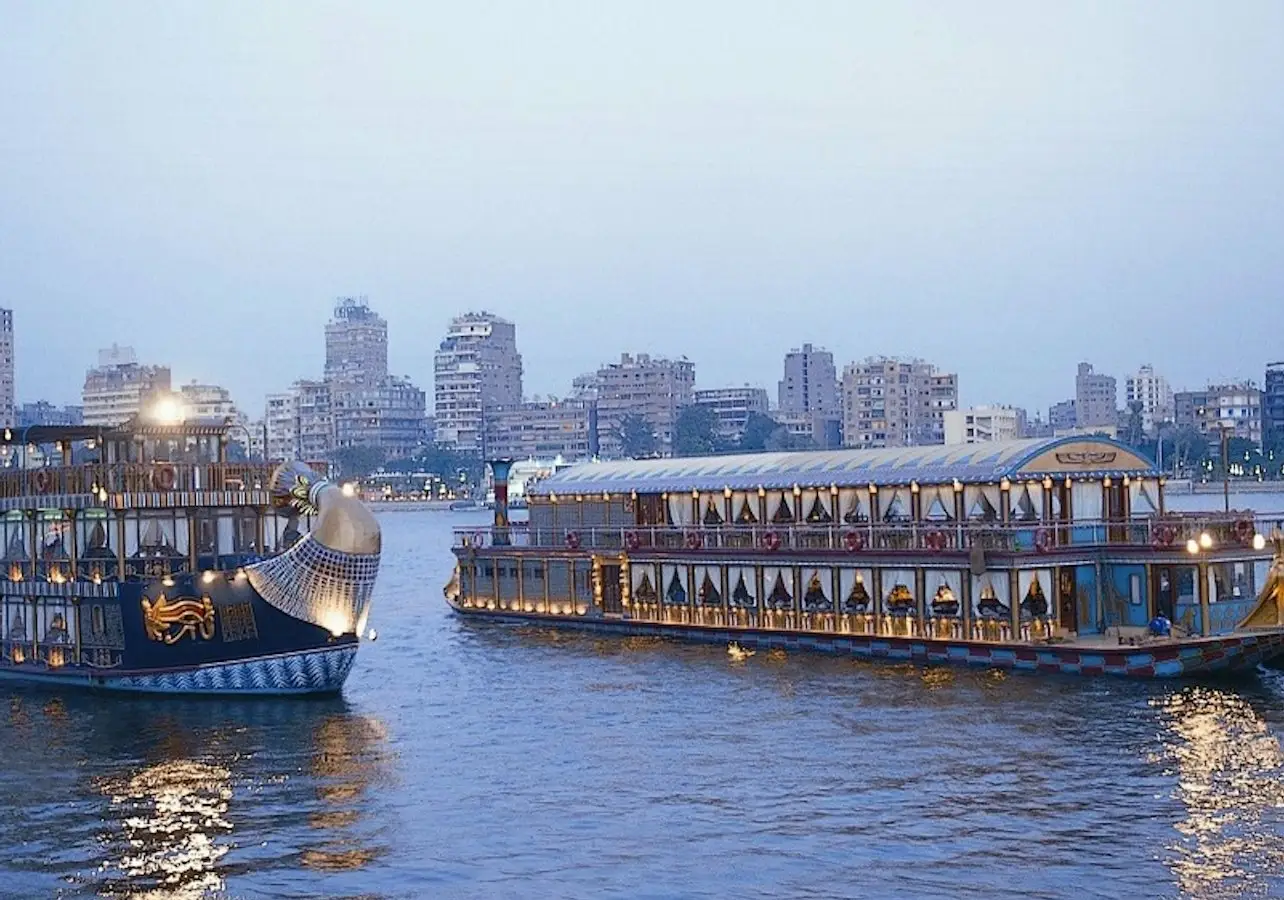 Two large passenger boats on a calm river with a city skyline and several high-rise buildings in the background during twilight.