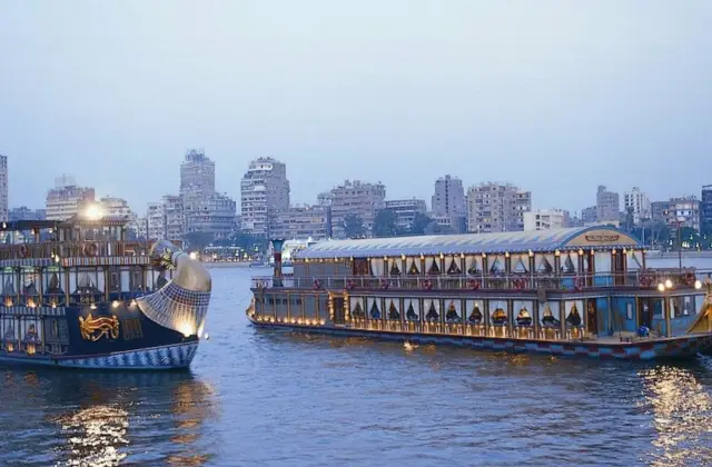 Two large passenger boats on a calm river with a city skyline and several high-rise buildings in the background during twilight.