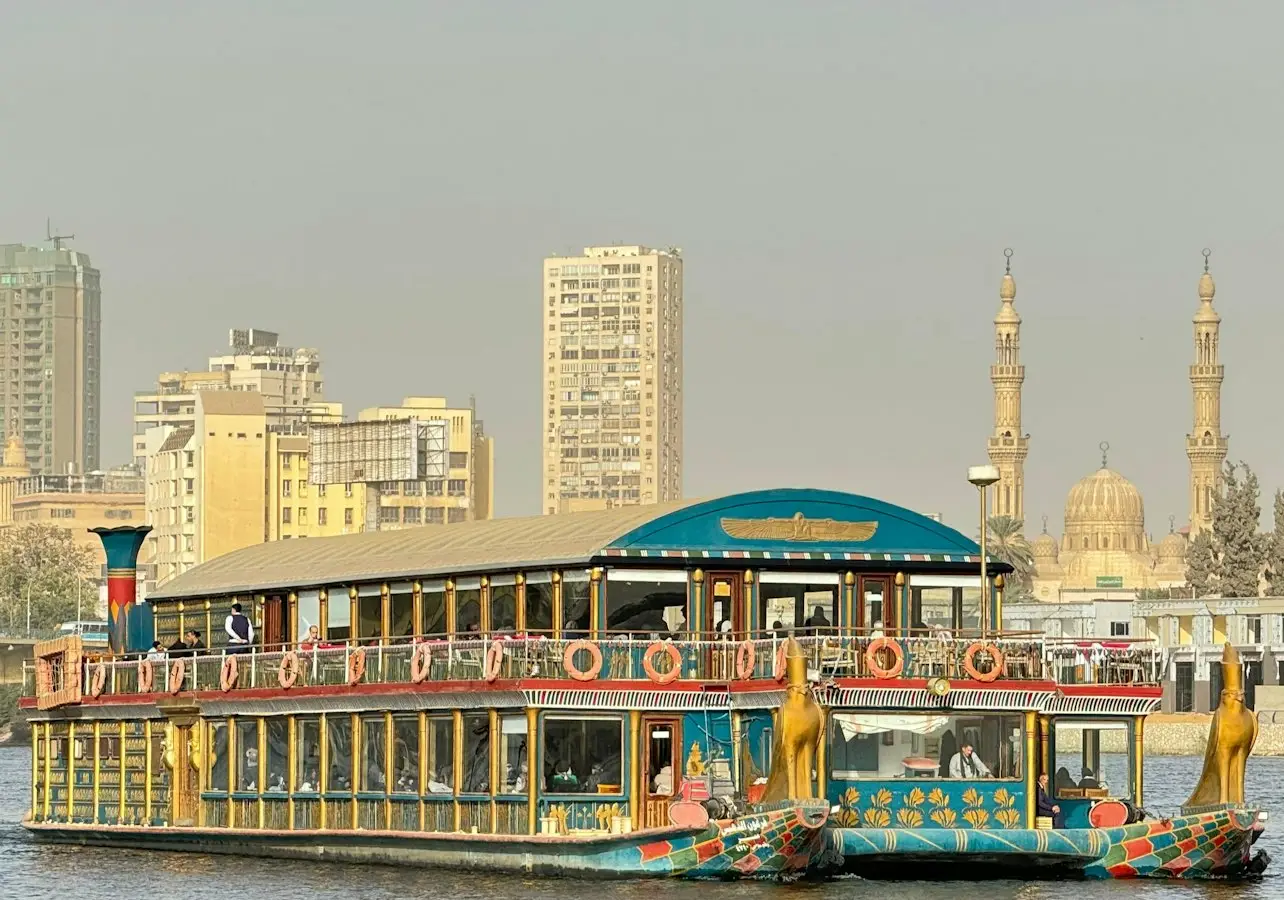 A riverboat styled with Egyptian motifs on a river, with a city skyline and two minarets in the background.