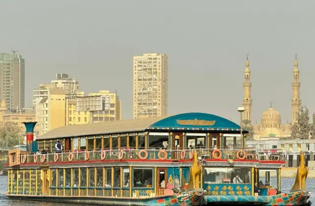 A riverboat styled with Egyptian motifs on a river, with a city skyline and two minarets in the background.