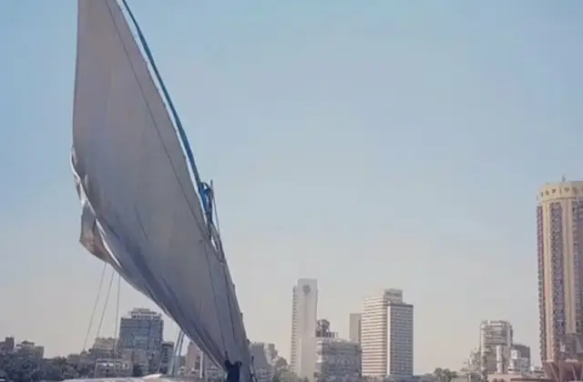 A sailboat with a large sail on a calm river, with a city skyline featuring tall buildings in the background under a clear sky.