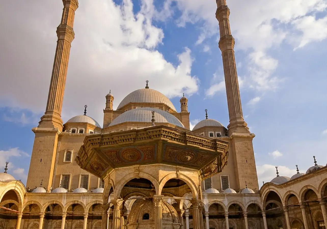 A large mosque courtyard with two tall minarets, a central ornate structure, domes, and a blue sky with clouds.