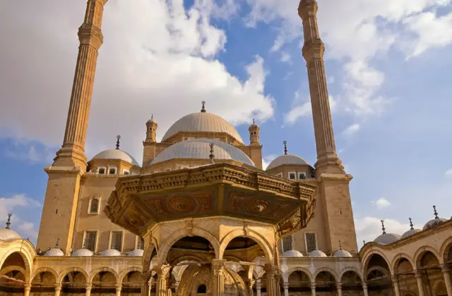 A large mosque courtyard with two tall minarets, a central ornate structure, domes, and a blue sky with clouds.