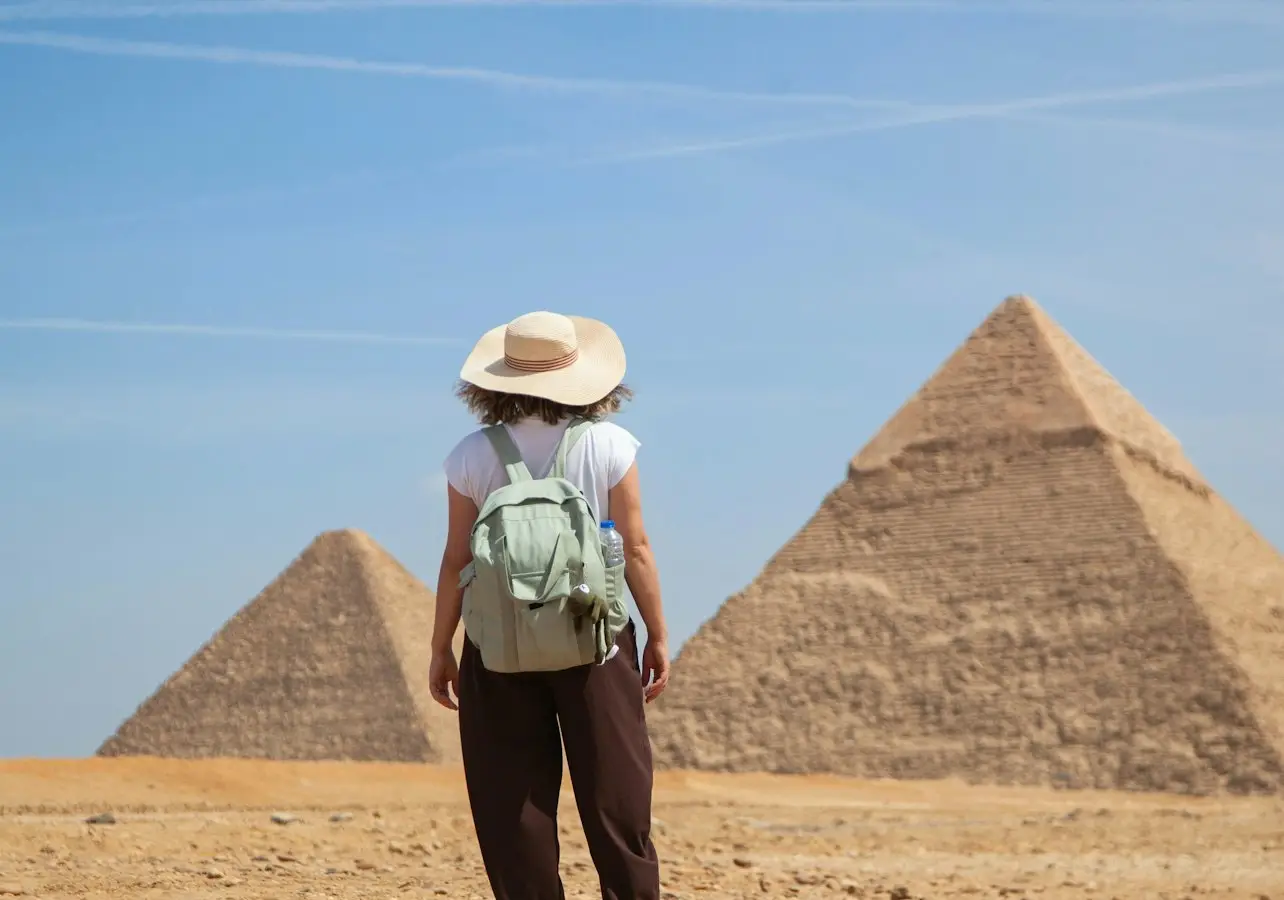 A person wearing a hat and backpack stands in front of two pyramids under a blue sky.