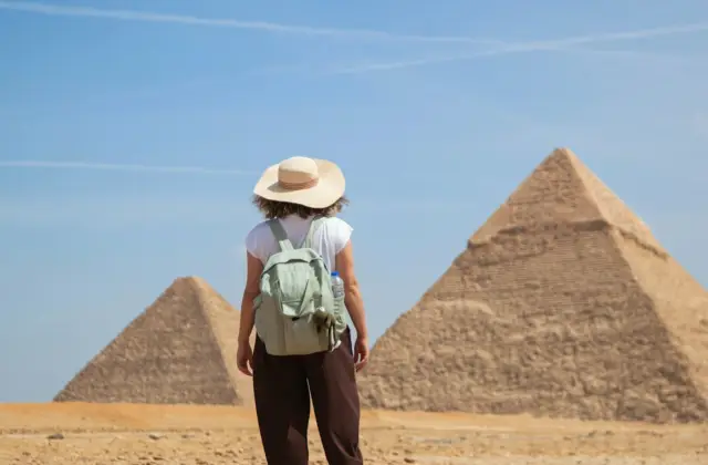 A person wearing a hat and backpack stands in front of two pyramids under a blue sky.