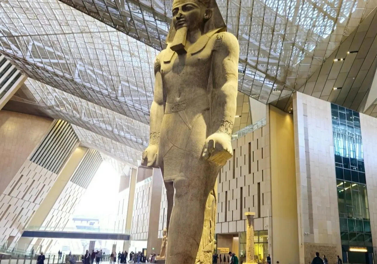 A large statue of an ancient Egyptian figure stands in a modern indoor space with a high ceiling and glass walls.