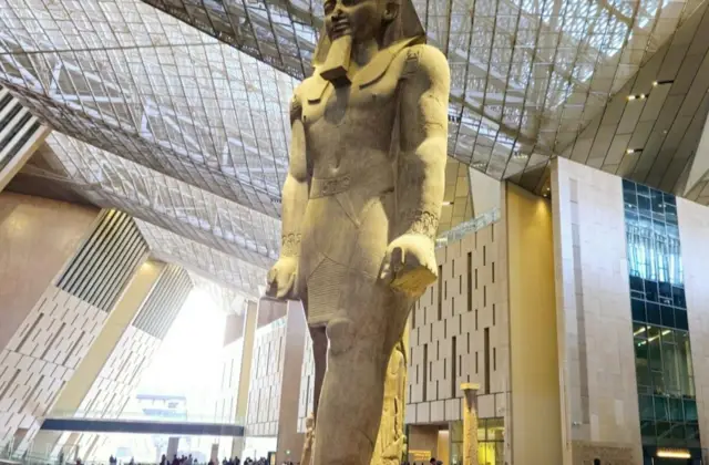 A large statue of an ancient Egyptian figure stands in a modern indoor space with a high ceiling and glass walls.