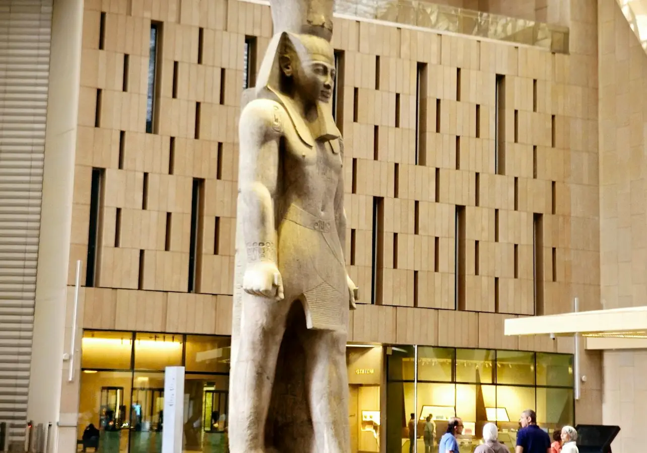 A large ancient Egyptian statue displayed in a modern museum interior, with people viewing it nearby.