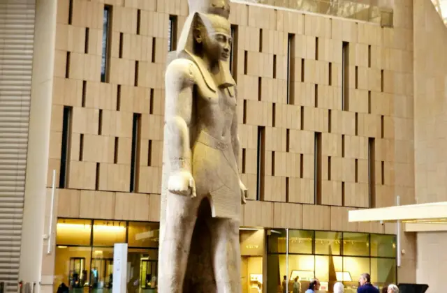 A large ancient Egyptian statue displayed in a modern museum interior, with people viewing it nearby.