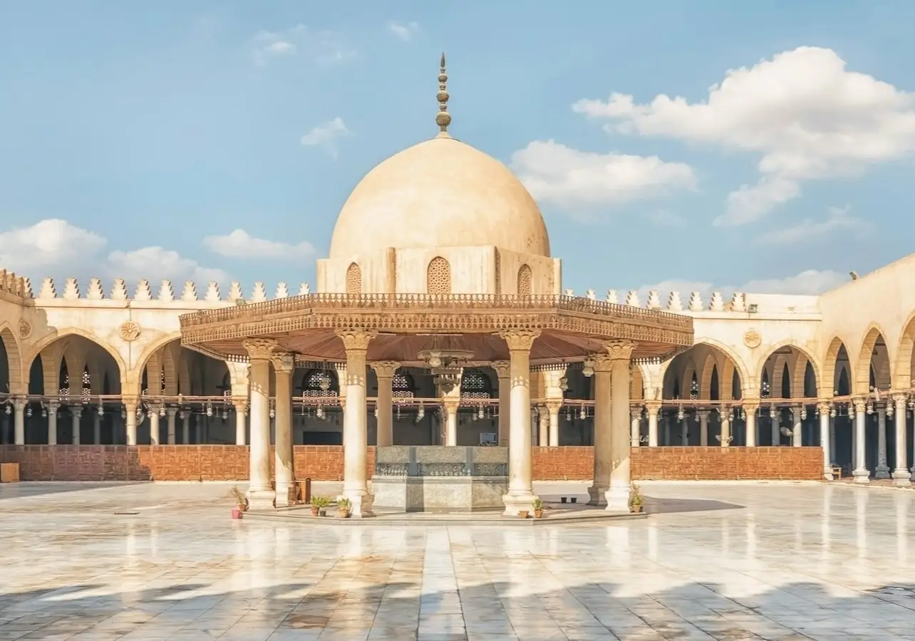 A mosque courtyard with a central dome structure, surrounded by arches and columns, under a blue sky with a few clouds.