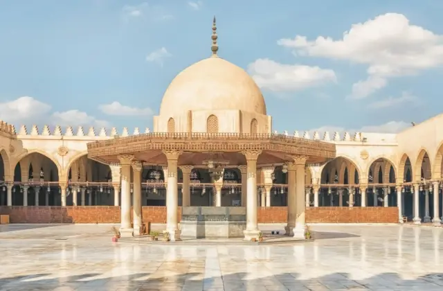A mosque courtyard with a central dome structure, surrounded by arches and columns, under a blue sky with a few clouds.