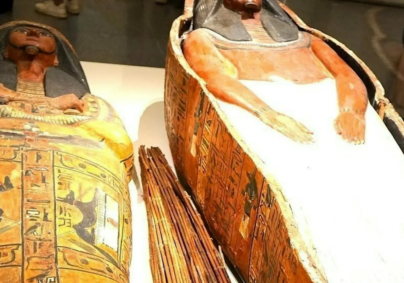 Two ancient Egyptian sarcophagi, one decorated with hieroglyphs and the other partially covered in a white shroud, displayed in a museum.
