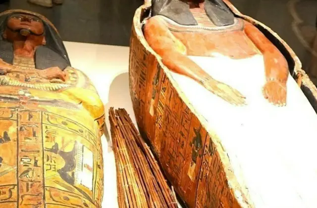 Two ancient Egyptian sarcophagi, one decorated with hieroglyphs and the other partially covered in a white shroud, displayed in a museum.