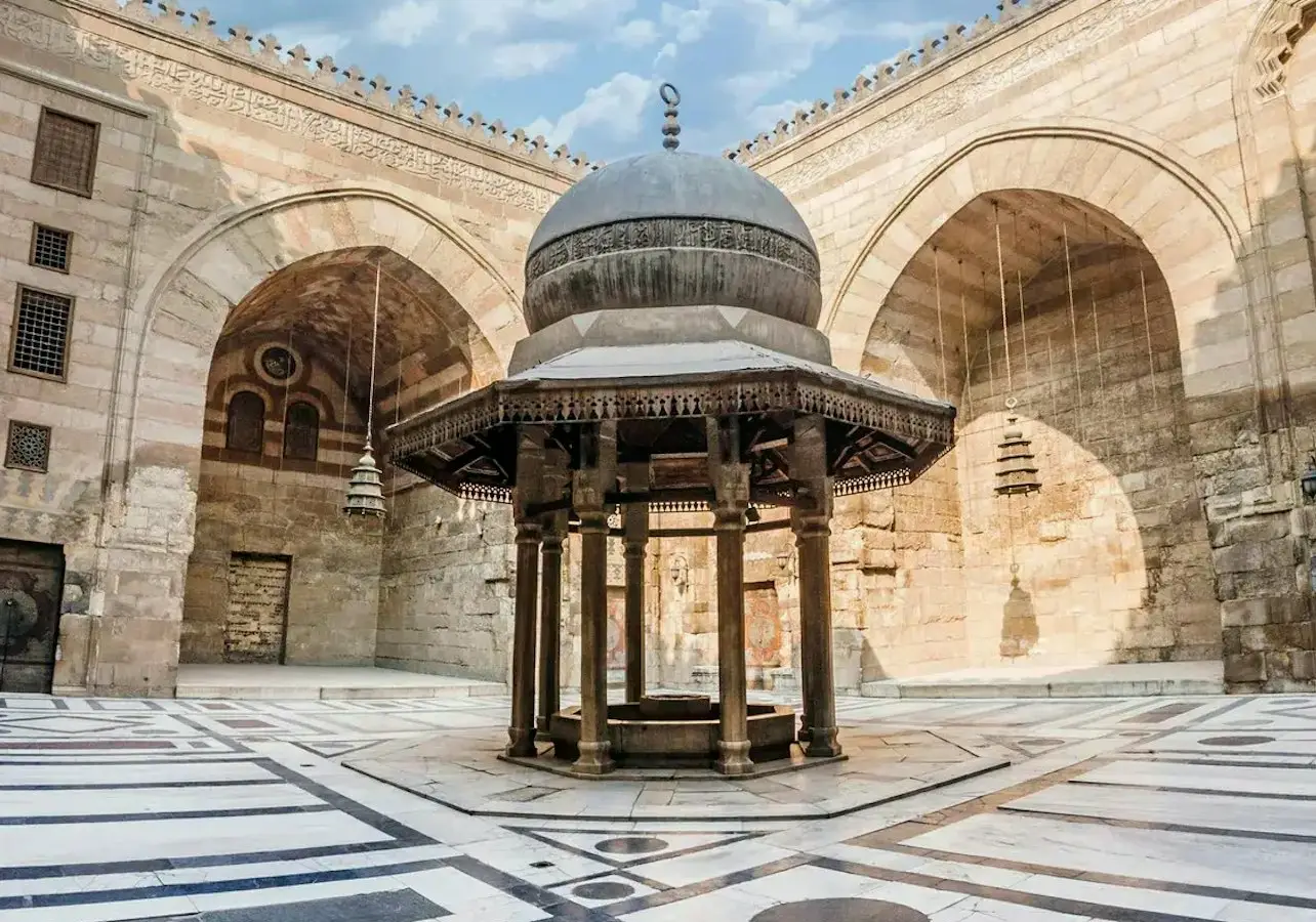 Courtyard of a historical building with a domed fountain surrounded by arches and patterned stone flooring under a blue sky.