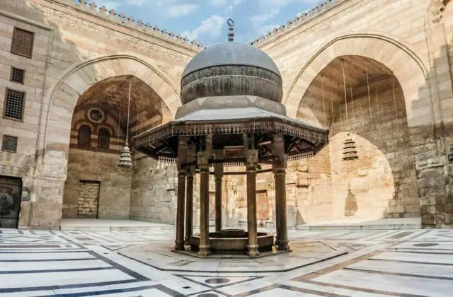 Courtyard of a historical building with a domed fountain surrounded by arches and patterned stone flooring under a blue sky.