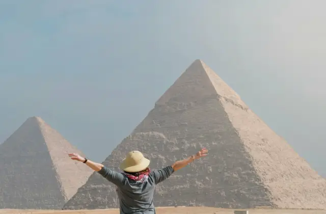 Person with outstretched arms facing the pyramids of Giza on a clear day.