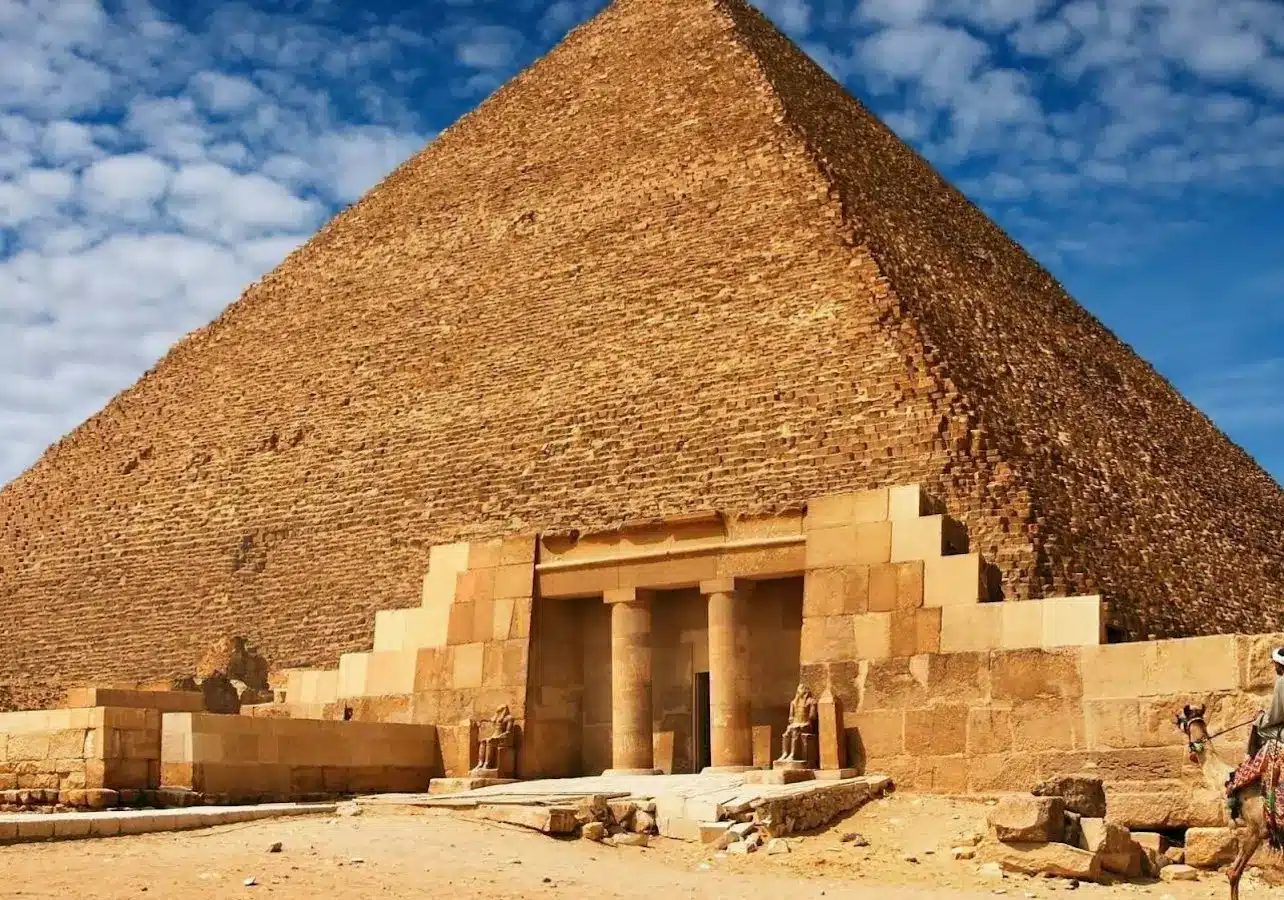 The Great Pyramid of Giza under a blue sky with scattered clouds, showing its entrance and surrounding sandy ground.