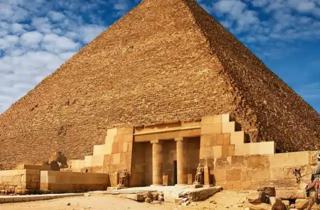 The Great Pyramid of Giza under a blue sky with scattered clouds, showing its entrance and surrounding sandy ground.