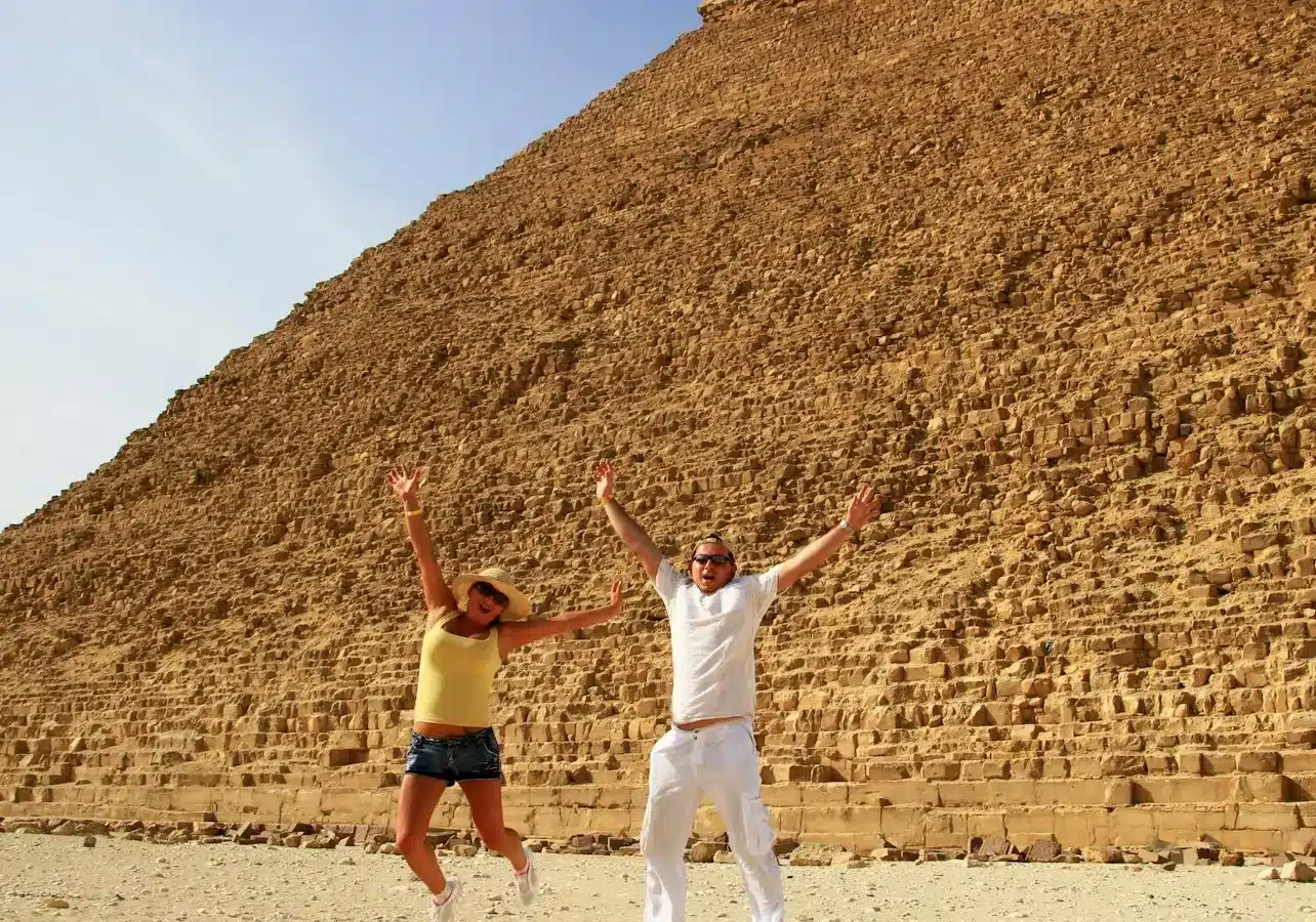 Two people jumping in front of the Great Pyramid of Giza, clear sky in the background.