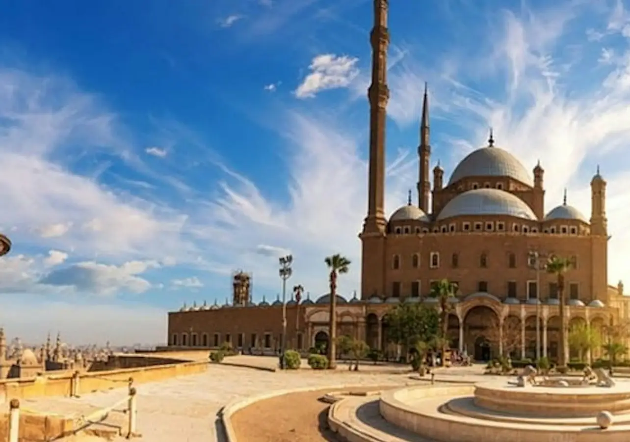 A grand mosque with multiple domes and minarets under a bright blue sky with scattered clouds, surrounded by a courtyard.