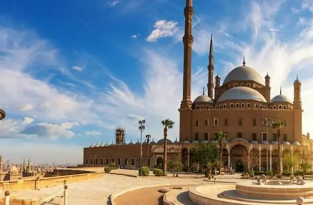 A grand mosque with multiple domes and minarets under a bright blue sky with scattered clouds, surrounded by a courtyard.