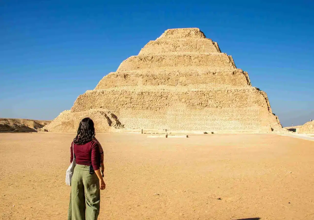 A person walking towards the Step Pyramid of Djoser under a clear blue sky in a desert setting.